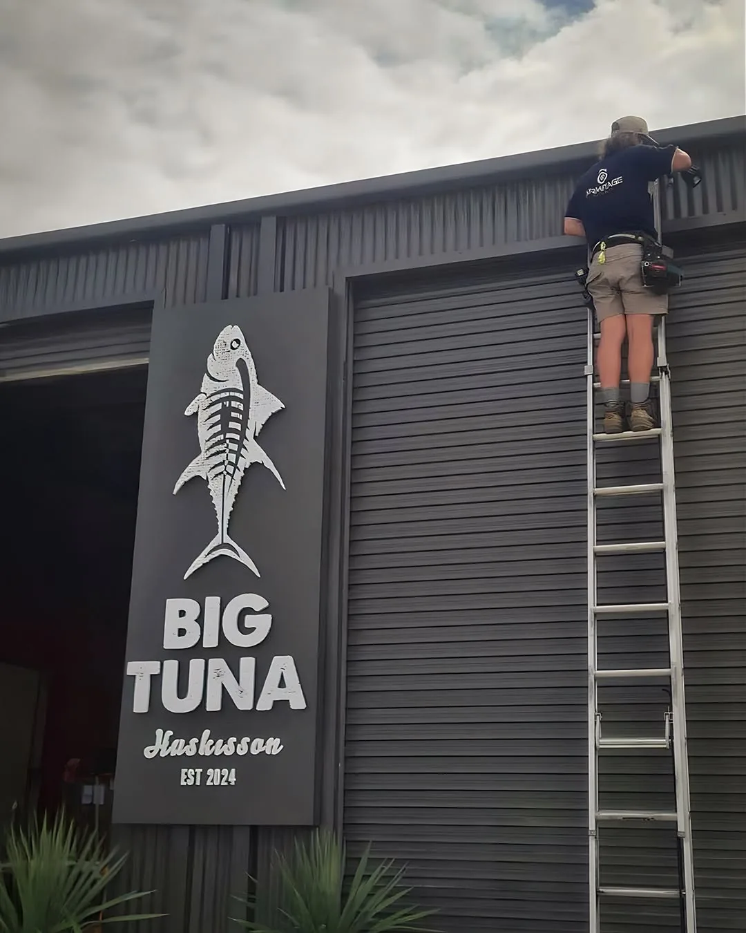 An Armitage Security technician on an extension ladder fitting a camera to a wall above the Big Tuna sign in Huskisson