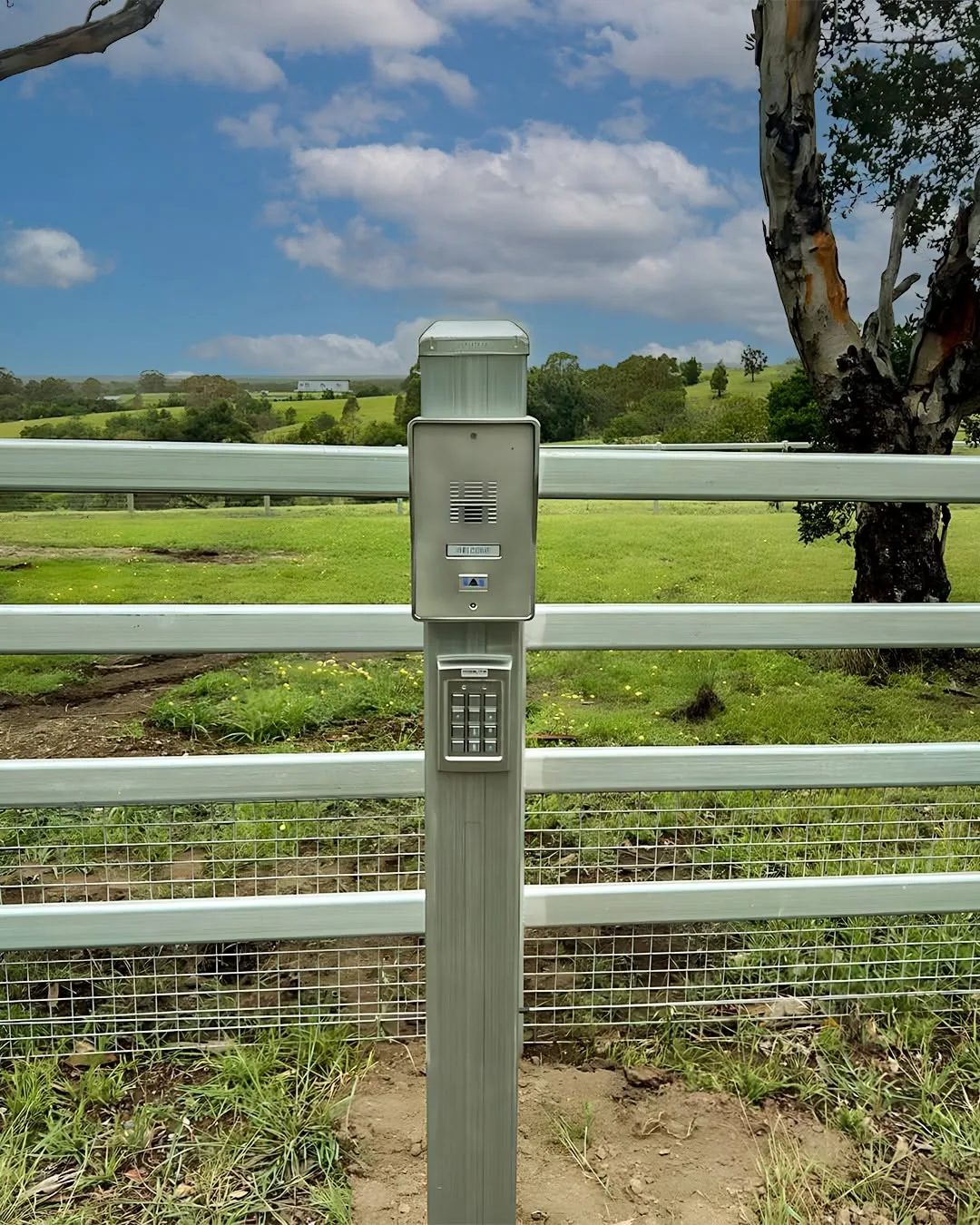 An intercom keypad post at a white post and rail fence with paddocks behind