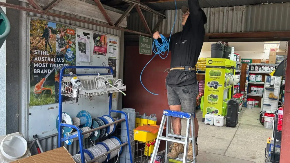 An Armitage Security technician on a ladder running cable through the roof trusses of a rural hardware store