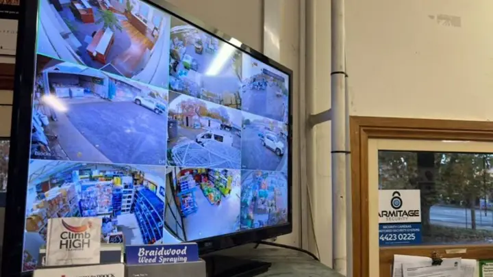 A wall monitor showing a nine camera CCTV grid in a rural store, the Armitage Security sign beside it