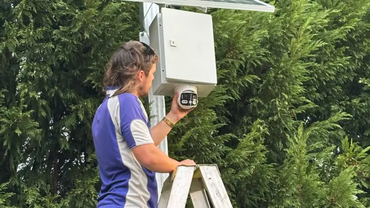 An Armitage Security technician on a ladder fitting a solar powered camera in a hedge