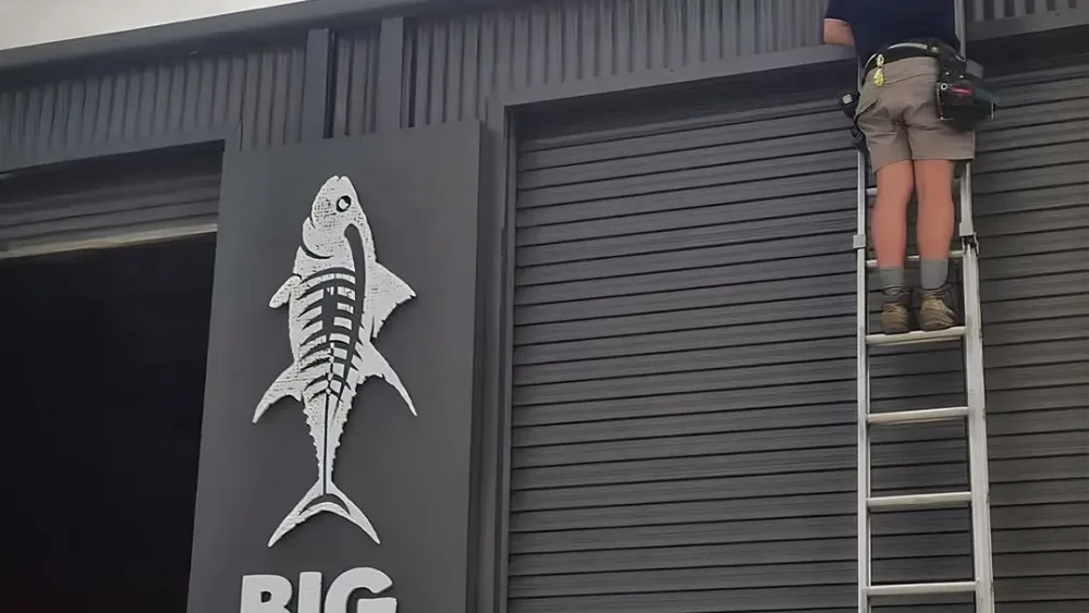An Armitage Security technician on an extension ladder fitting a camera to a wall above the Big Tuna sign in Huskisson