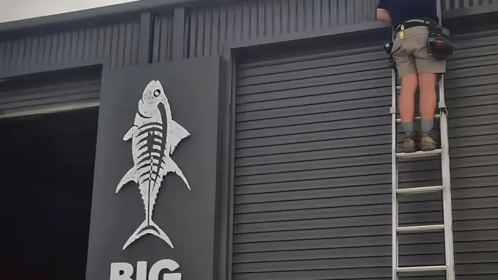 An Armitage Security technician on an extension ladder fitting a camera to a wall above the Big Tuna sign in Huskisson